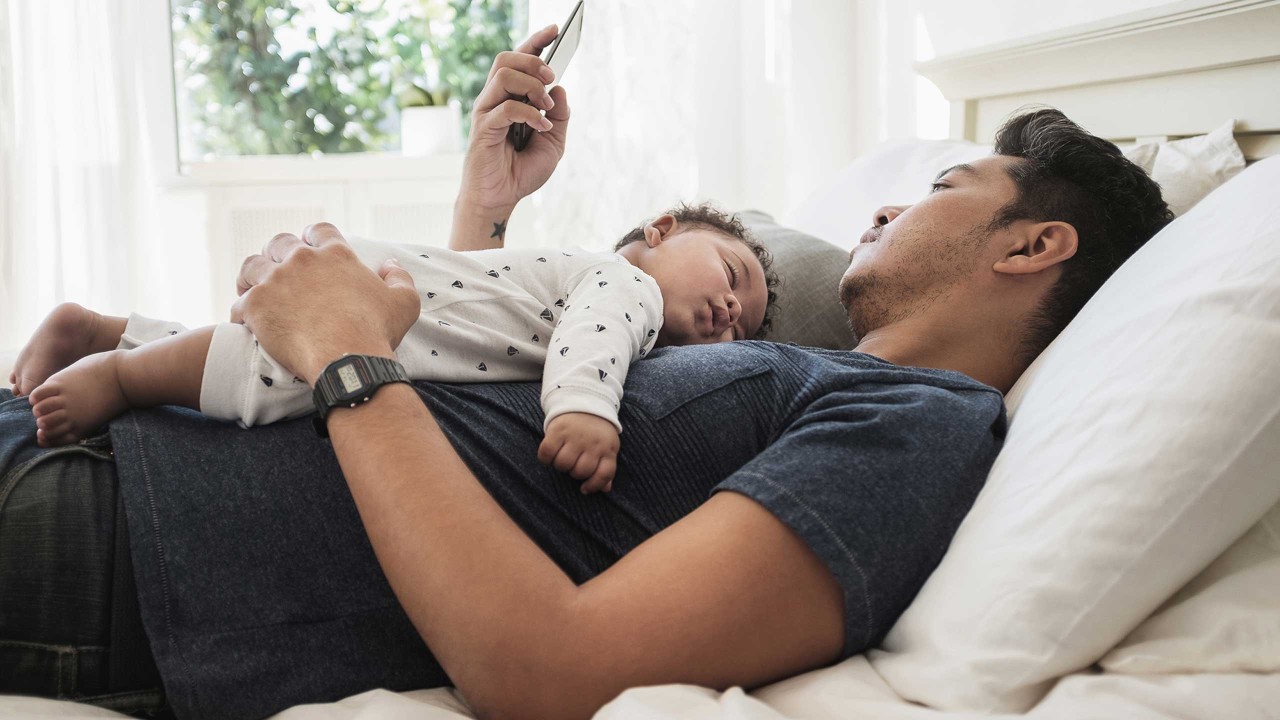 a father is using mobile phone on bed while his baby sleeping soundly on his chest; image used for HSBC Mauritius relief measures page.
