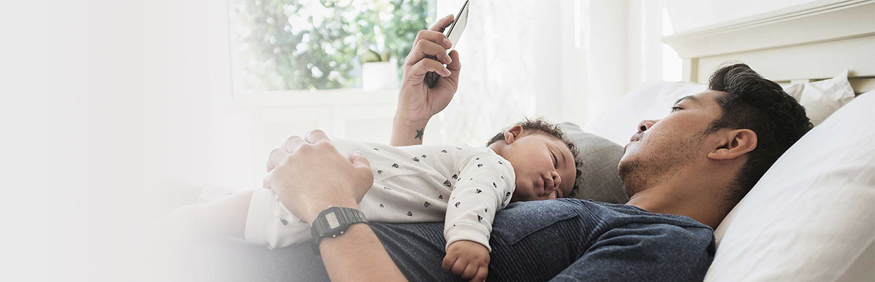 a father is using mobile phone on bed while his baby sleeping soundly on his chest; image used for HSBC Mauritius relief measures page.